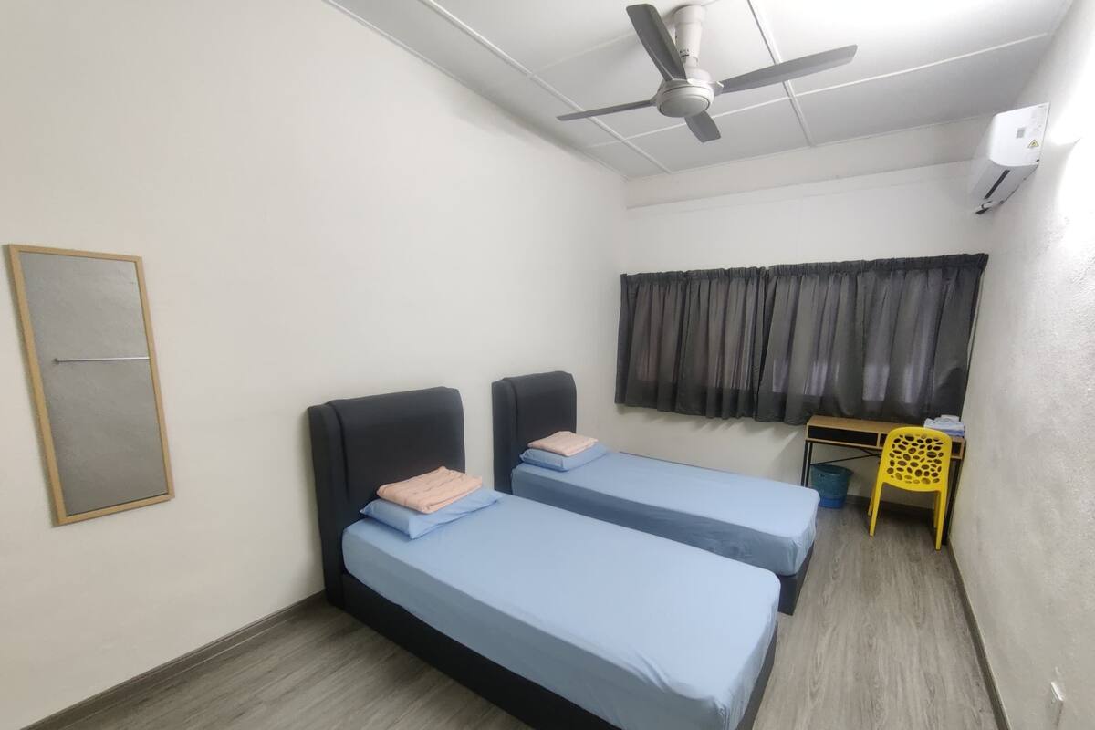 A room with two single beds, a ceiling fan, a window with curtains, and a small table with a chair.