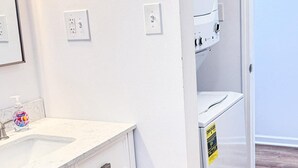 Casa de banho com toucador branco, pia e espelho. Uma porta dá para outro espaço.