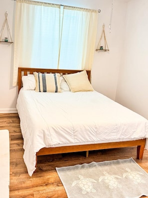 A bedroom with a wooden bed, white bedding, a wooden floor, a window with curtains, and a small rug.