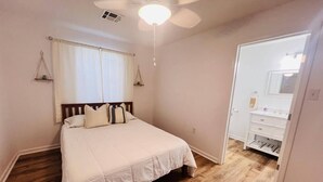 Quarto com cama, ventilador de teto, janela com cortinas, piso de madeira e banheiro com pia e espelho.