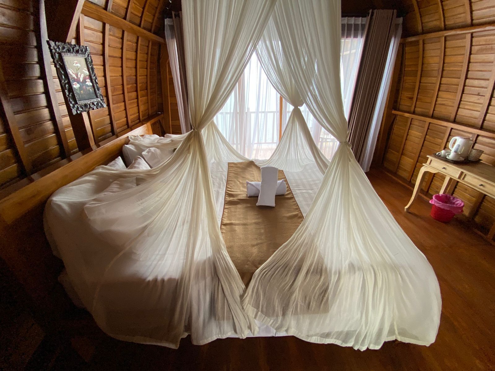 A room with a bed covered in white sheets, a wooden headboard, and a canopy bed with sheer white curtains. There is a small table with a kettle and a pink trash bin.