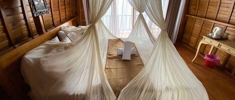 A room with a bed covered in white sheets, a wooden headboard, and a canopy bed with sheer white curtains. There is a small table with a kettle and a pink trash bin.