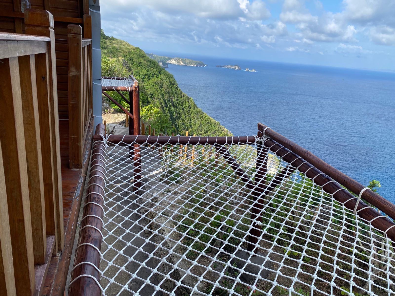 A wooden balcony with a mesh safety railing overlooking a coastal landscape with cliffs and the sea.