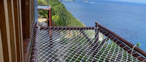 Un balcón de madera con barandilla de seguridad de malla que da a un paisaje costero con acantilados y el mar.