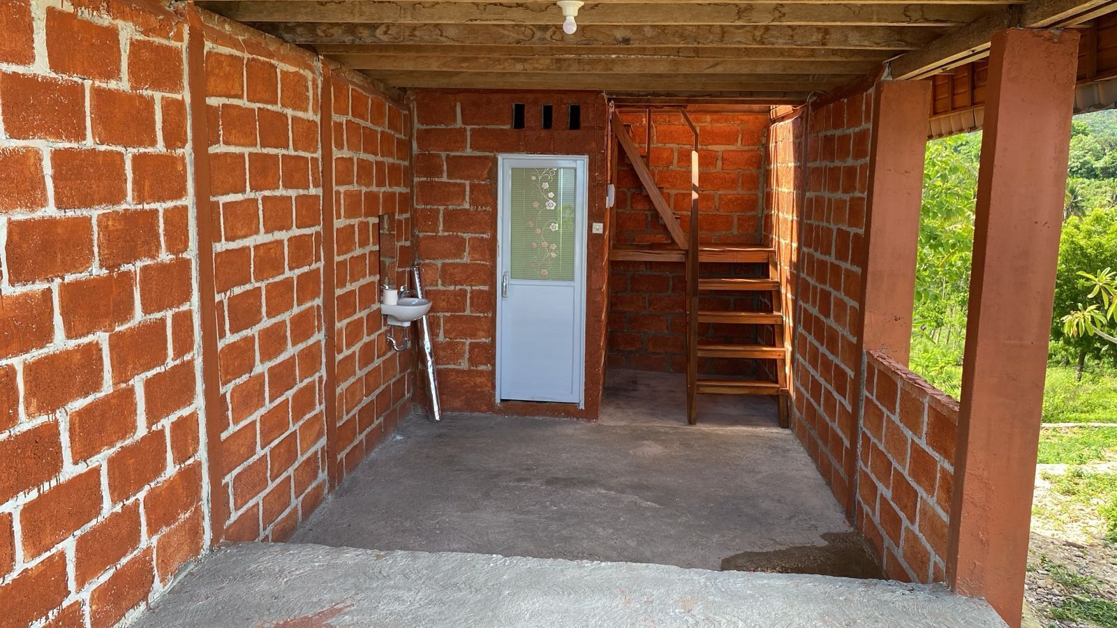 Une petite pièce en briques, fermée, avec une porte blanche, une échelle en bois et un sol en béton.