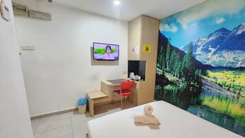 A hotel room with a bed, a desk, a chair, a TV, and a large wall mural of a mountain landscape.