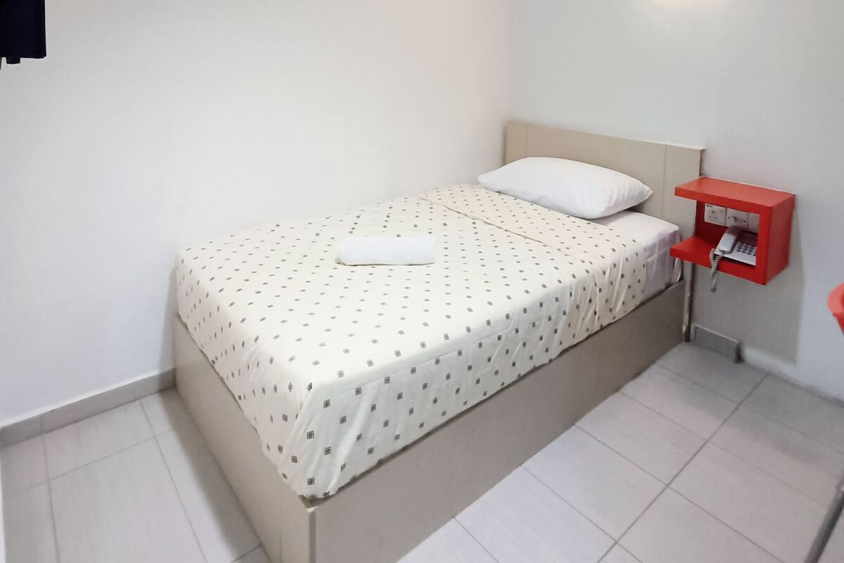 Una habitación pequeña con cama individual, colcha blanca, mesita de noche roja con teléfono y una lámpara de pared.
