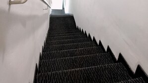 Uma escadaria com degraus pretos e paredes brancas, conduzindo a uma porta com a placa “KELUAR”.