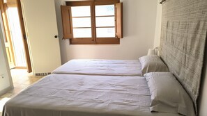 Two single beds with white mattresses and pillows, a wooden window with shutters, and a tiled floor.