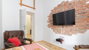 A room with a brick wall, a mounted TV, a wooden table with a checkered tablecloth, a leather armchair, and a red pillow.