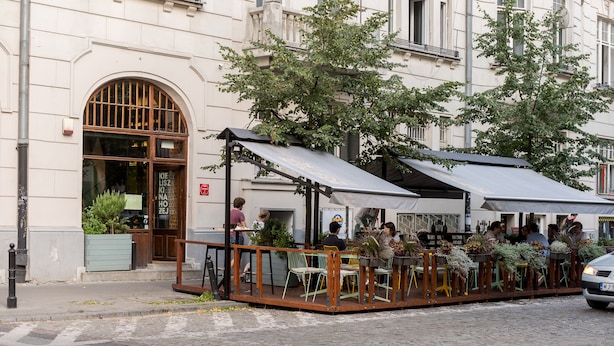 An outdoor café with tables and chairs, surrounded by a wooden fence, in front of a building with large windows and a door. - City Centre All In One Apartment (Warszawa)
