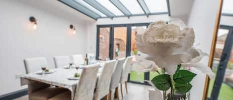 A dining area with a table set for four, a white rose in a vase, and a view of the outside through large windows.