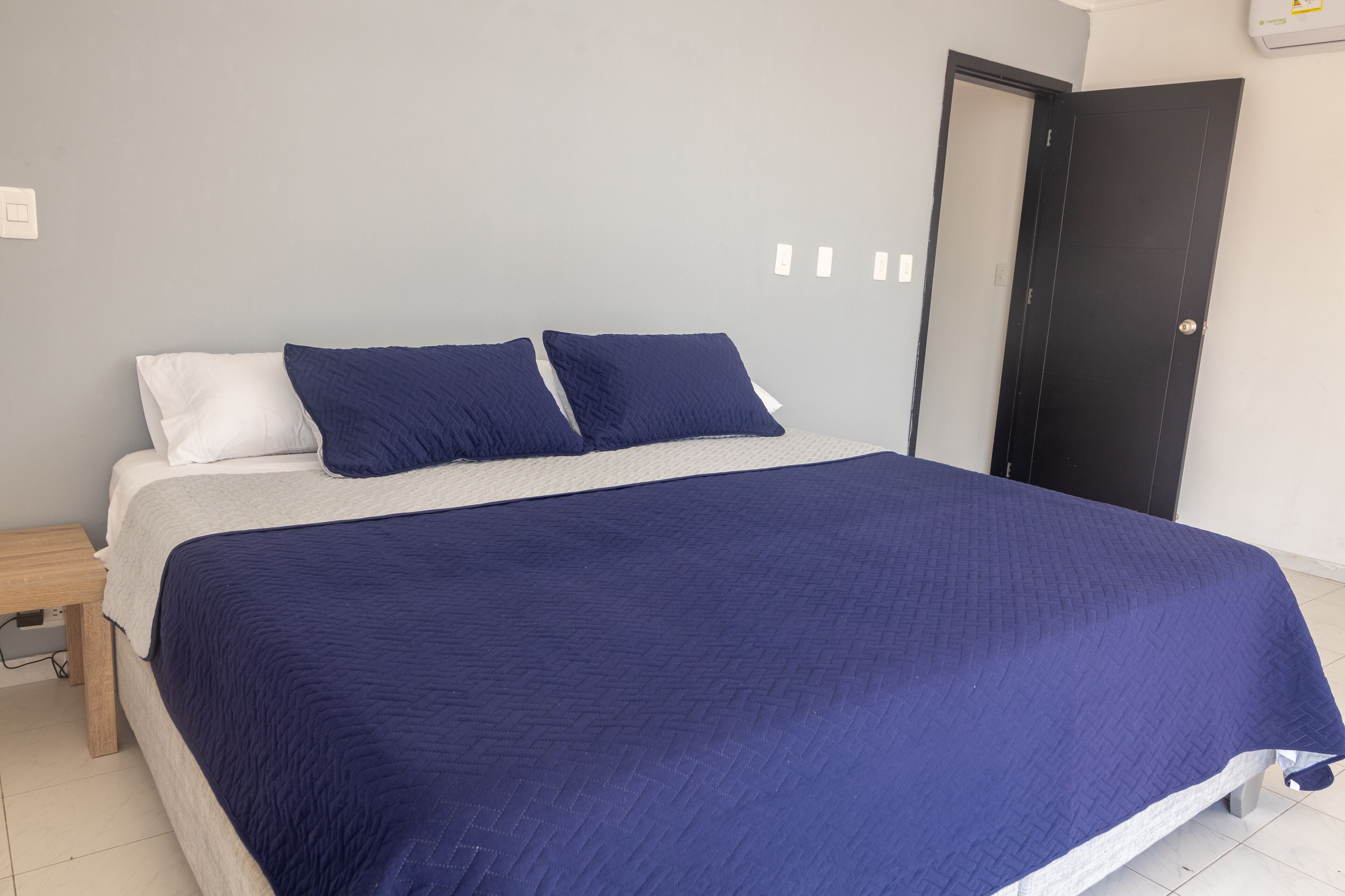 A bed with a white and blue quilt, a wooden nightstand, and a black door.
