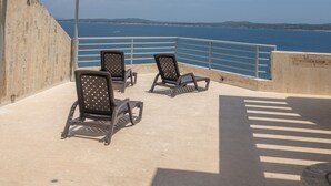 A terrace with two lounge chairs facing a body of water, a railing, and a clear sky.