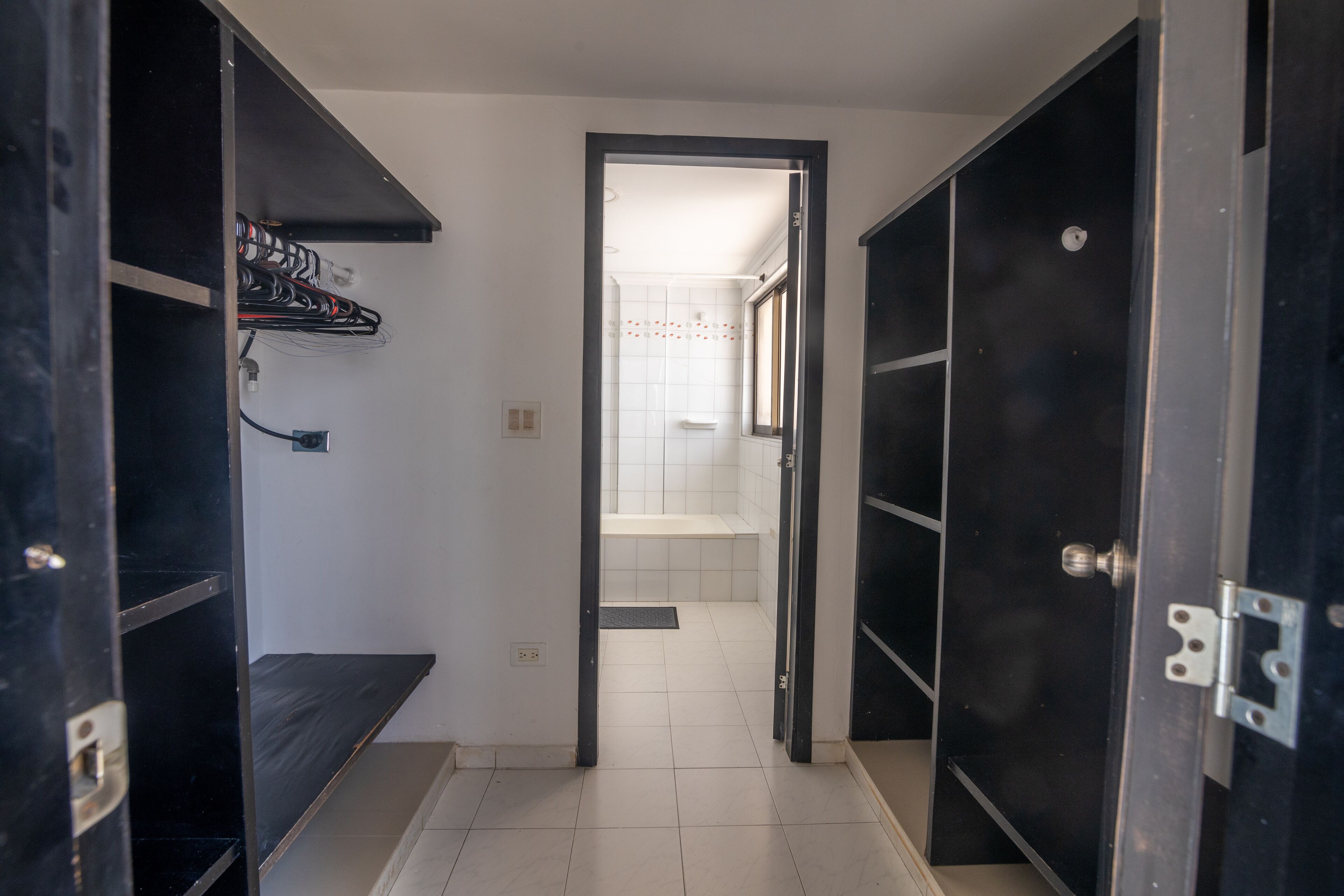 A narrow hallway with a door leading to a bathroom. The hallway has a tiled floor and a shelf with a few items on it.