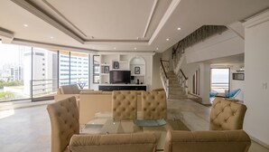 A spacious living area with a glass coffee table, beige sofas, a flat-screen TV, and a staircase leading to an upper floor.