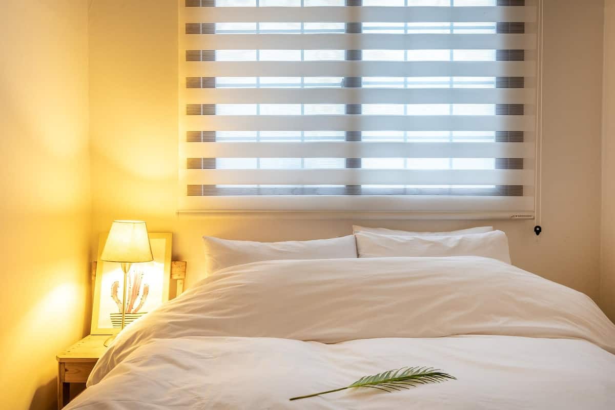 A neatly made bed with white linens, a bedside lamp, and a window with blinds.