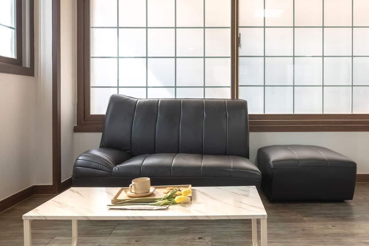 A modern living room with a dark leather sofa and ottoman, a marble coffee table with a cup and a book, and a large window with a grid pattern.