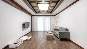 A modern hotel room with a flat-screen TV mounted on the wall, a grey sofa, a small wooden coffee table, and a pet bed. - Geoje Blueberry Pet Pension (Geoje)