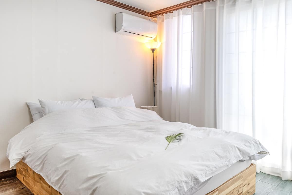A bedroom with a wooden bed frame, white bedding, a single green leaf decoration, a wall-mounted air conditioner, and sheer curtains allowing natural light.