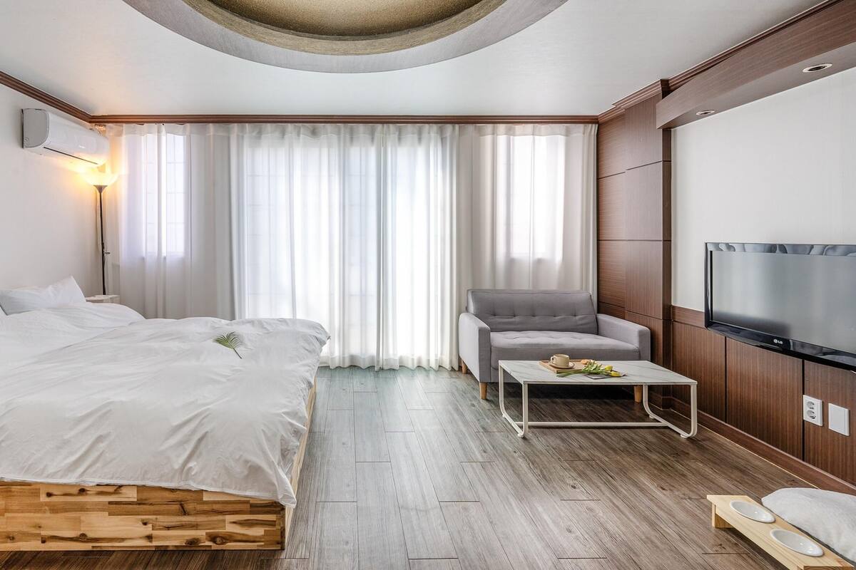 A modern hotel room with a large bed, a grey armchair, a small table with a cup and flowers, a flat-screen TV, and a wooden floor.