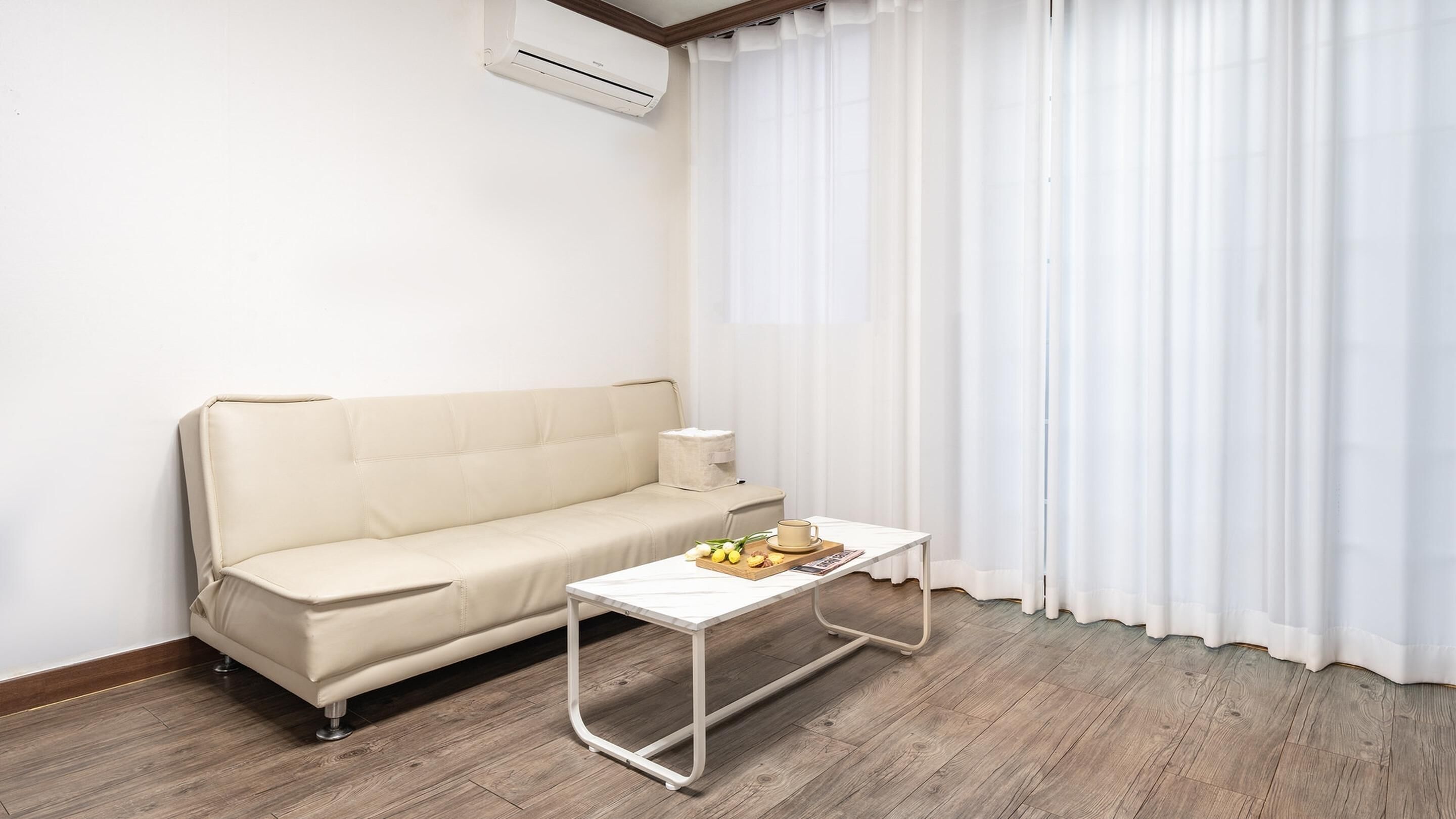 Un salón minimalista con un sofá beige, una mesita con una bandeja de limones y una taza, y una ventana con cortinas blancas.