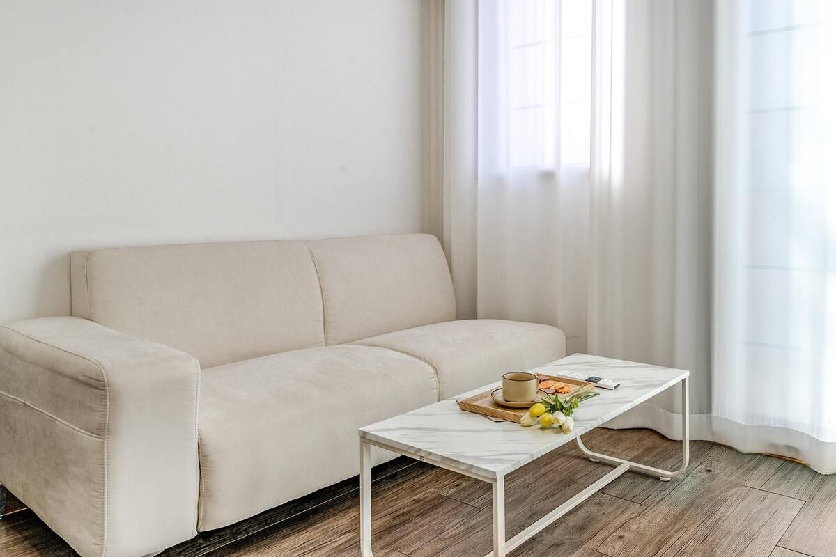 A minimalist living room with a beige sofa, a white coffee table with a tray of snacks, and a window with sheer curtains.