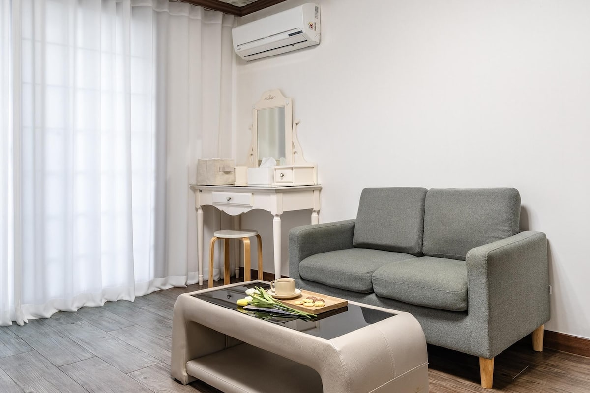 A modern living room with a grey sofa, a small round table with a tray of cookies and a cup, a white vanity with a mirror, and a window with white curtains.