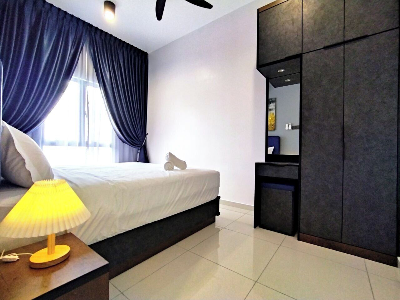 A modern hotel room with a bed, a nightstand with a lamp, a wardrobe, and a large window with curtains.