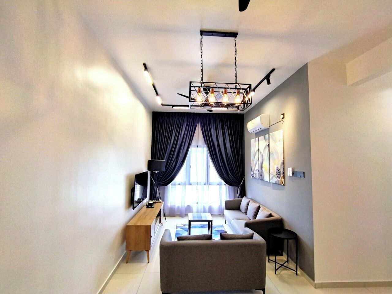A modern living room with a sofa, coffee table, and TV. There is a large window with curtains, a ceiling light fixture, and a painting on the wall.