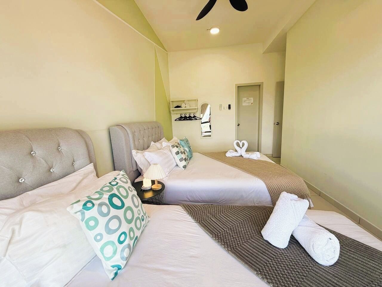 A hotel room with two beds, a ceiling fan, a small table with a lamp, and a door leading to another room.