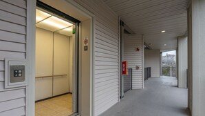 Un couloir avec une porte donnant sur un ascenseur, un détecteur d’incendie et une vue sur un bâtiment situé au loin.