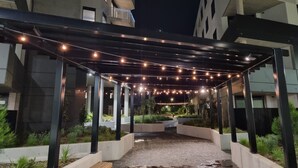 Un espace extérieur abrité, agrémenté de guirlandes lumineuses, de piliers en béton et d’un chemin pavé.