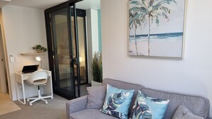 Un salon moderne avec un canapé gris, deux coussins à motifs, un bureau avec un ordinateur portable et une porte-fenêtre coulissante donnant sur un balcon.