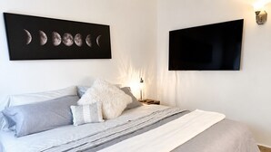Een slaapkamer met een bed, kussens, een dekbed en een aan de muur bevestigde televisie, evenals een kunstwerk met maanfasen in een lijst.