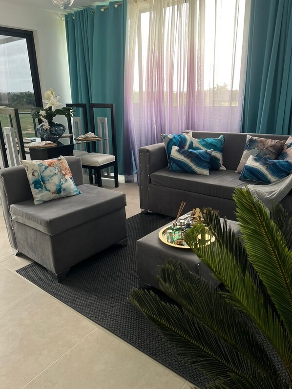 A modern living room with a grey sofa, a grey armchair, and a coffee table with a tray. There is a large window with blue curtains, a vase with flowers, and a view of the outdoors.