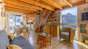 Een gezellig interieur van een chalet met houten plafond, een bank, een eettafel, een rieten stoel en een televisie.