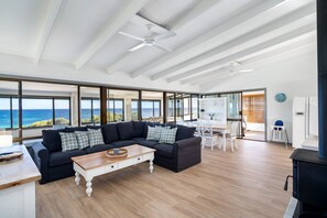 Een ruime zithoek met een grote, donkergrijze modulaire bank, een houten salontafel en uitzicht op de oceaan door grote ramen.