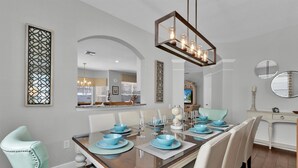 A dining area with a wooden table set for six, surrounded by white chairs with teal accents. - Windsor Hills Resort 6 Bedroom Private Pool+Spa Home (Kissimmee)