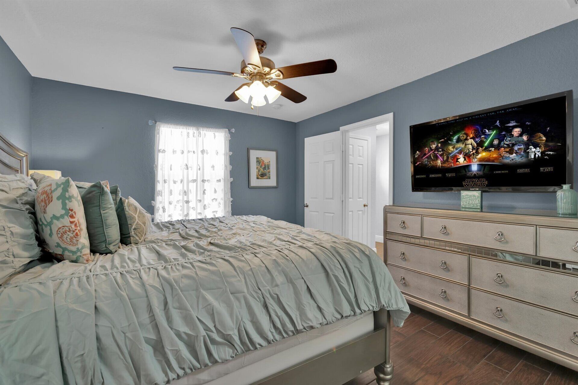 A bedroom with a bed, a ceiling fan, a dresser, and a TV displaying a movie poster.