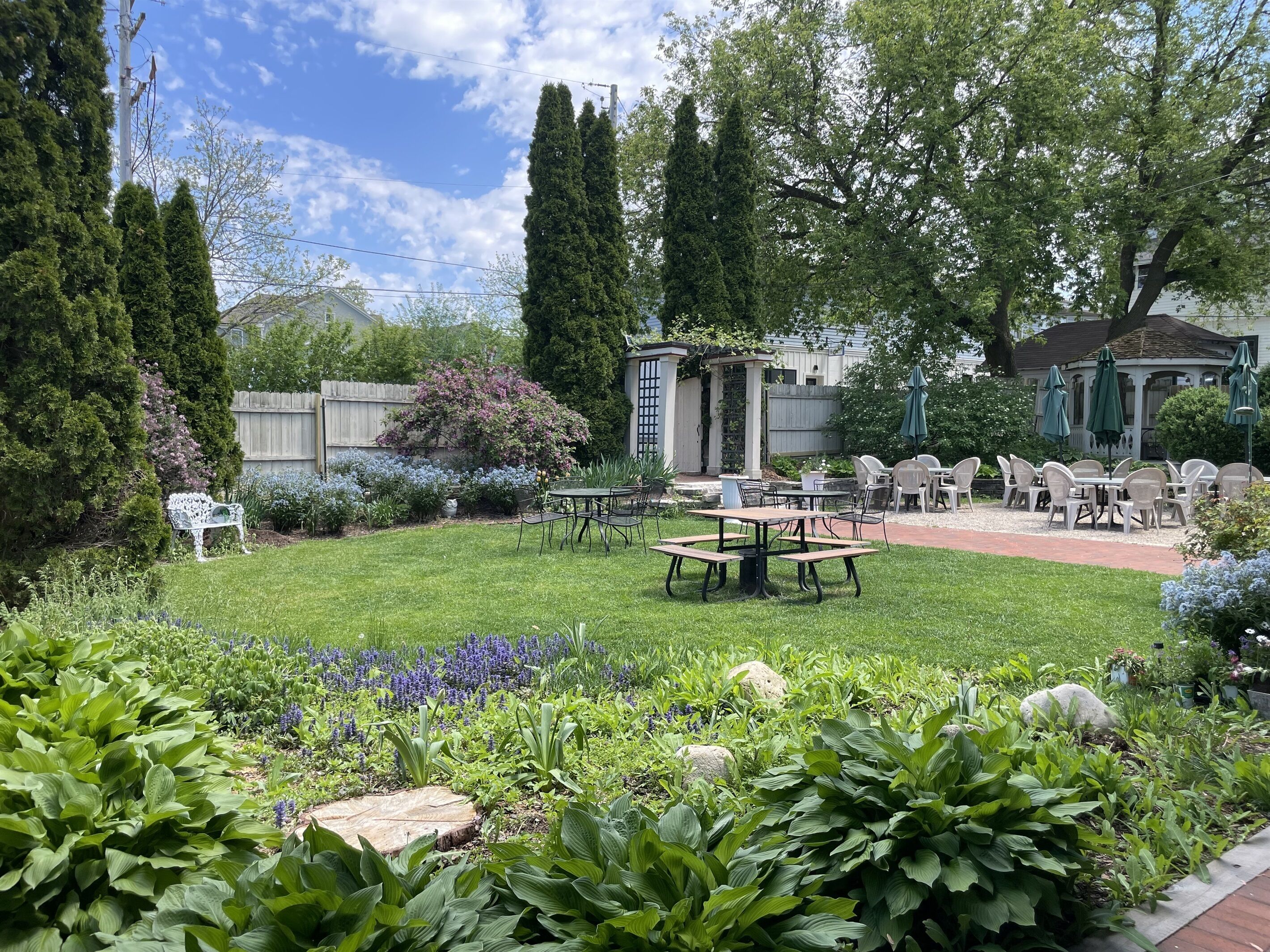 A well-maintained garden with a patio area featuring tables and chairs, surrounded by various plants and trees.