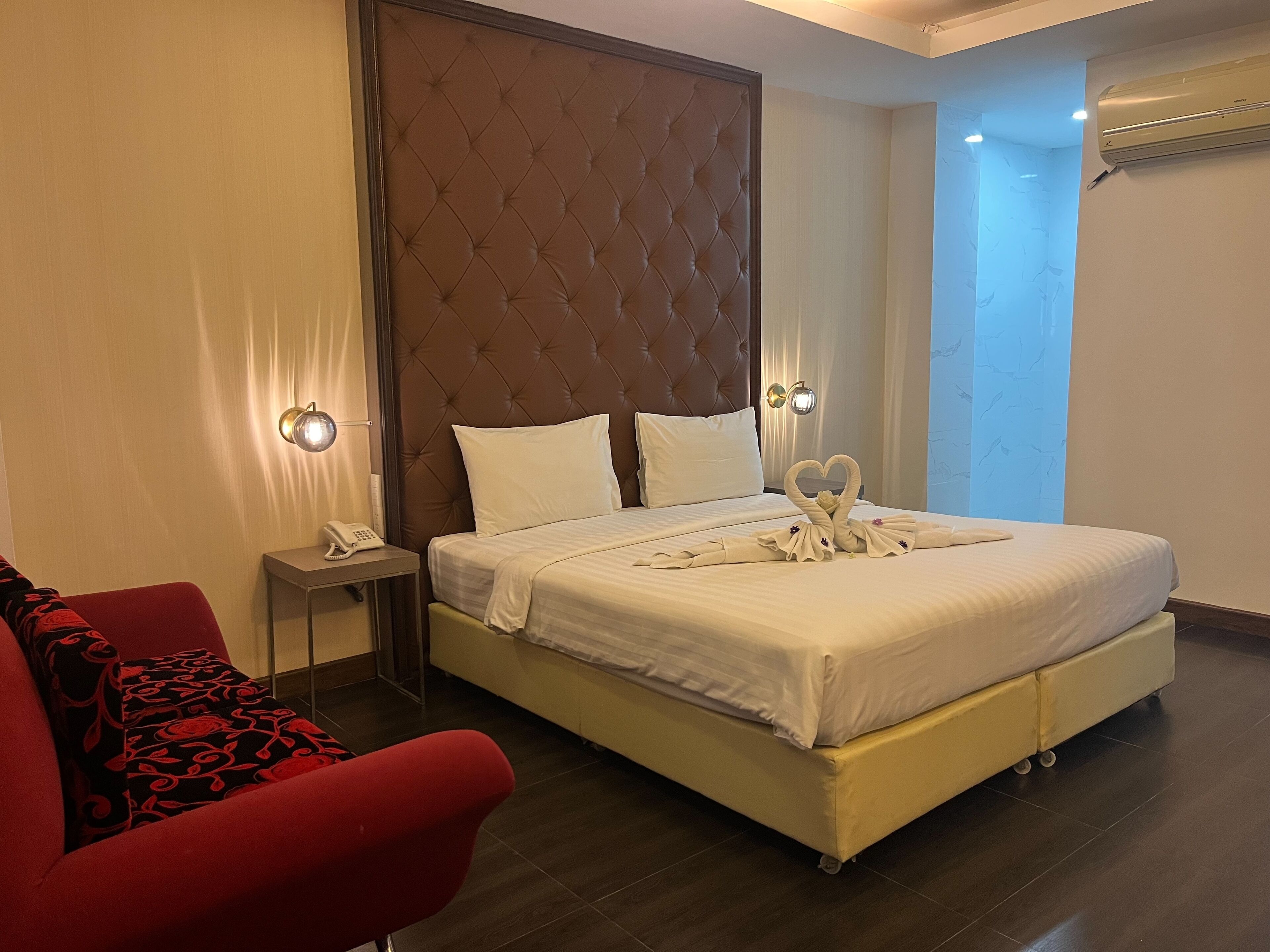 A hotel room with a large bed, a red armchair, a bedside table with a phone, and a wall-mounted air conditioner.