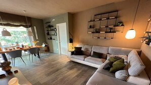 Een moderne woonkamer met een bank, eettafel en stoelen. Er is een boekenplank en een lamp aan de muur.
