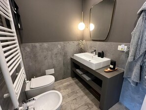 Een moderne badkamer met een witte wasbak, een toilet en een bidet. Boven de wasbak hangt een spiegel en aan de rechterkant een handdoekhouder.