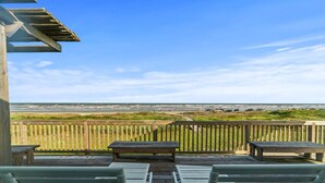 A wooden deck with white lounge chairs and a table, overlooking a beach with a wooden fence and grassy dunes.
