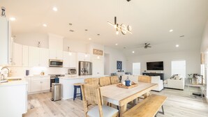 A spacious kitchen with white cabinets, a central island, and modern appliances. There is a dining area with wooden chairs and benches, and a living room with a sofa and a flat-screen TV. - Get ready to soak up the sun at Villa Caman! (Galveston)