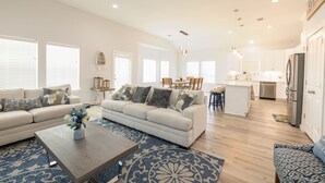 A spacious living room with a sofa, coffee table, and patterned rugs. A kitchen area with a refrigerator and dining table is visible in the background. - Get ready to soak up the sun at Villa Caman! (Galveston)