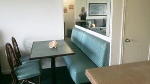 Et spiseområde med et bord og stole, en blå polstret bænk og et spejl der afspejler en stue med en sofa og en person.