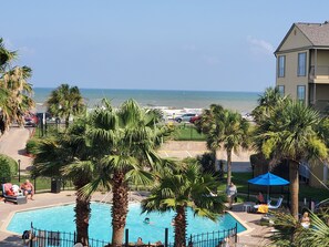A pool area with palm trees, a view of the beach, and a building with balconies. - Welcome to the Victorian, Dig your toes in the sand or enjoy a dip in the pool! (Galveston)