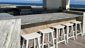 An outdoor kitchen with a view of the ocean, featuring a grill, bar seating, and a stone countertop. - Good Day Sunshine - Emerald 712 (Galveston)